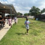 Children running on a grassy field beside a covered patio where adults sit at picnic tables for a casual event.