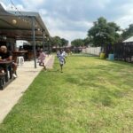 Outdoor cafe area with people sitting at long benches under a covered patio; kids run on the grass beside them.