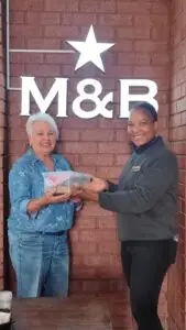 Two smiling women exchange a clear plastic lunch container in front of a brick wall with a bright M&B sign and star above them (donation/food handoff).
