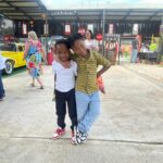 Two boys pose with arms around each other, smiling at the camera at an outdoor fair; a yellow vintage car and vendor stalls visible behind them.