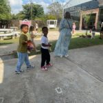 Children carrying small bags walk along a concrete path in a sunny outdoor courtyard.