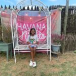 Girl sits on a white ornate garden swing in front of a pink HAVANA banner, outdoors with planters and a wooden fence nearby.