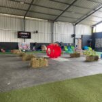 Indoor children's play area with hay bales, colorful seating, and a large red circular play structure in the center.