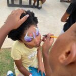 A child sits with eyes closed while a painter applies a unicorn face design with a purple and white horn and pink flowers on the forehead.