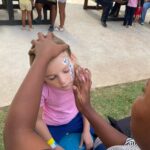 A young girl sits with eyes closed as a face painter adds blue flower designs to her forehead at an outdoor event with kids and picnic tables in the background.