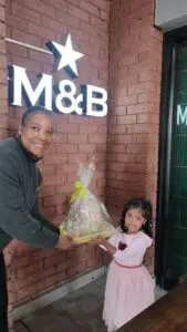 Woman in a dark jacket hands a gift basket wrapped in cellophane with a yellow ribbon to a smiling little girl in a pink dress, outside a brick storefront with an M&B sign and star logo.