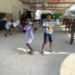 Two boys pose with snacks in a decorated open-air play area; colorful tassels hang from the ceiling and other kids play in the background.
