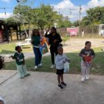 Children holding gift bags at an outdoor community event on a sunny day under string lights.