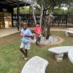 Two children walk across a grassy yard, each holding snack bags, near a circular stone garden and outdoor tables at a park-like area.