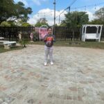 Smiling girl stands on a circular cobblestone patio in a yard, holding a red-and-blue toy toward the camera. A wooden fence, string lights, and a white garden swing are in the background.