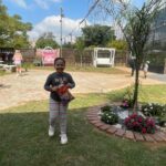 Girl in striped pants holding a snack bag, running toward the camera in a sunny yard with a circular brick flower bed garnished with flowers.
