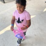 Young girl in a pink shirt carries a pink drawstring bag with small blue items, outdoors.