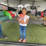 Young girl wearing bunny ears and a white top holds a wrapped gift at an Easter-themed indoor play area on artificial grass.