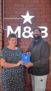 Two adults smile while exchanging a blue gift bag in front of a brick wall with a bright M18B sign and star above them.