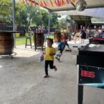 Two kids run with bags under a festively decorated outdoor shelter, pink/orange tassel garlands overhead.
