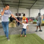 Adult man guiding a girl in a white hat as they walk on a small grassy mat in an indoor play space.