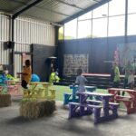 Indoor community event with pastel-painted picnic tables, hay bales, and a stage with balloon decorations and a bunny backdrop on the right.