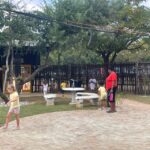 Child-friendly outdoor courtyard with stone tables and string lights; a woman in red supervises several kids nearby.