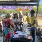 People gather around a table at an outdoor event under a roof, with a black tablecloth, baskets, and papers; a colorful Easter banner with rabbits and flowers hangs above.”,