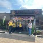 Group of volunteers in yellow shirts standing under a Happy Easter banner at The Boma Cafe in Sandton on a sunny day.