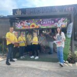 Group of people in yellow shirts stand outside a booth with a bright Happy Easter banner and bunny imagery.