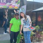 Two women wearing bunny ears pose for a photo outside a shop with an Easter banner showing a white bunny, smiling and holding hands; one wears a bright green shirt and the other a white shirt with blue pants, holding a colorful box of treats.