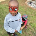 Young boy with Spider-Man face paint standing on grass, wearing a gray sweatshirt and black sneakers; another child wearing a colorful hat sits nearby outdoors.