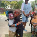 Two kids with Spider-Man face paint hug each other at an outdoor park party, a caregiver smiling in denim behind them.
