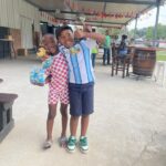 Two smiling boys stand together at an outdoor party; one in red‑and‑white checkered outfit holds snacks while the other in a blue and white striped shirt gives a thumbs up.