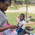 Woman applying colorful butterfly face paint to a young girl outdoors, with paint supplies on a table nearby.