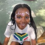 Smiling girl with braided hair and a white shirt featuring a South Africa flag heart, face painted with pink flowers, outdoors at a playground park.