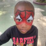 Young boy with Spider-Man face paint outdoors on a playground, wearing a black Garfield shirt with cartoon characters.
